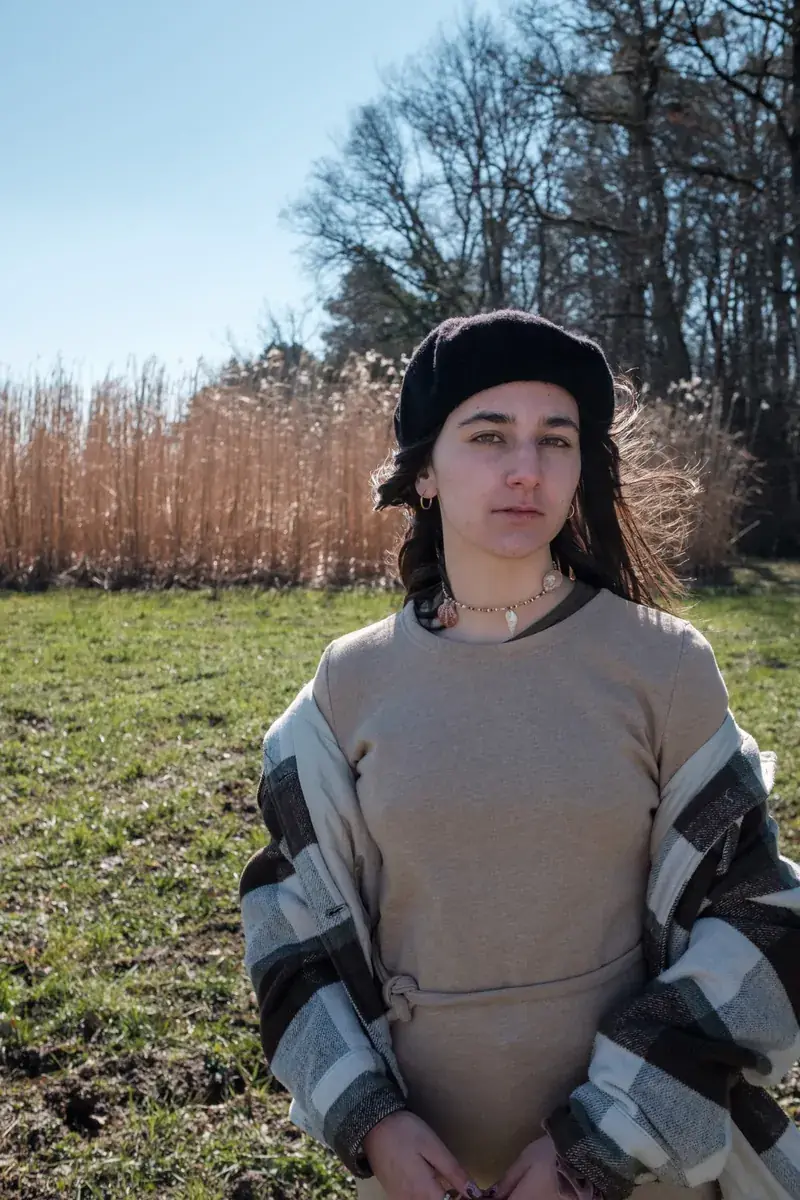 Une mannequin en béret noir et pull beige, dans un champ bordé de roseaux