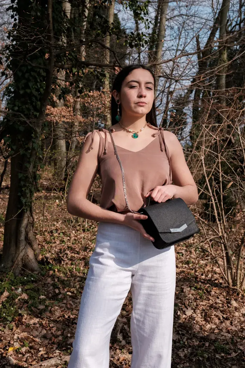 Une mannequin en top marron et pantalon blanc, un sac noir à la main, en forêt