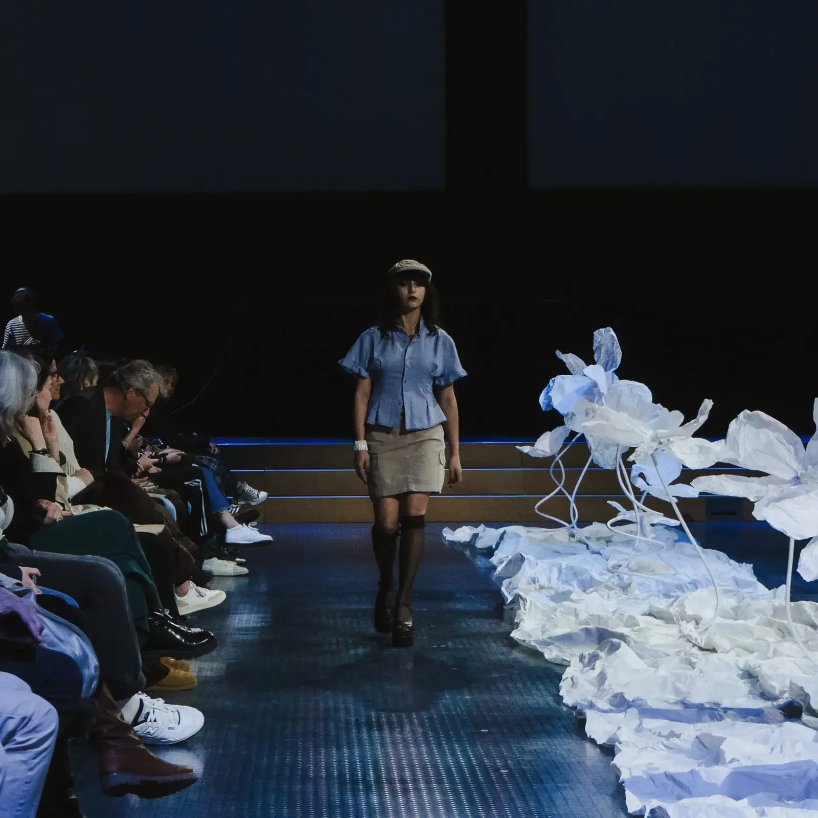 Une mannequin en chemise bleu clair et casquette défile à côté de grandes fleurs en papier blanc, devant le public de l'Ethical Fashion Show