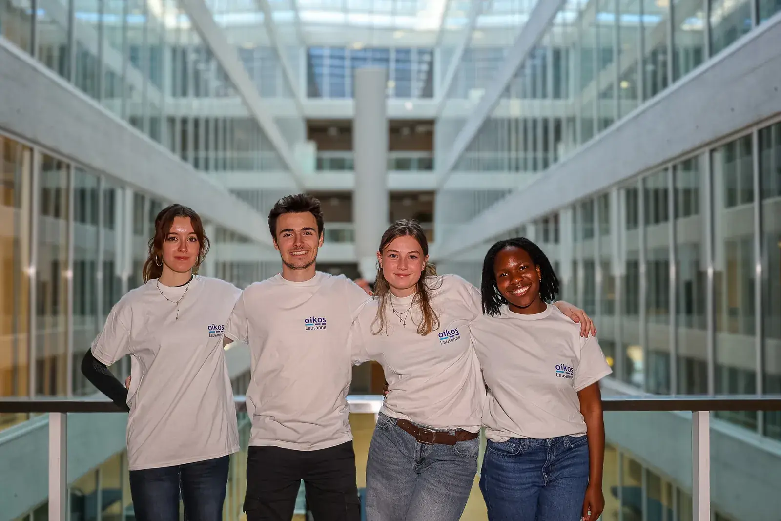 Valentine Krafft, Sename Bahun-Wilson, Eve Bouvier et Ugo Ortolini, le comité du printemps 2026, en t-shirt oikos sur une passerelle d'un bâtiment vitré du campus