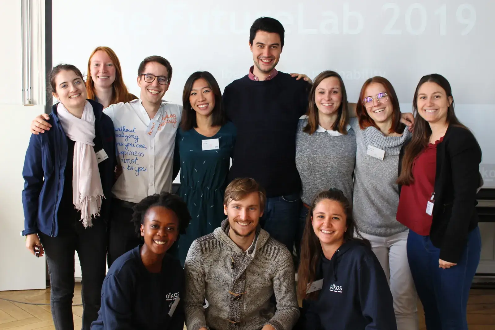 Une douzaine de membres d'oikos, plusieurs en sweat oikos, posent devant l'écran du FutureLab 2019